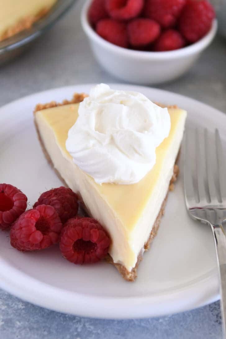 Top-down view of a slice of lemon pie with a dollop of whipped cream on a white plate, with fresh raspberries on the side.