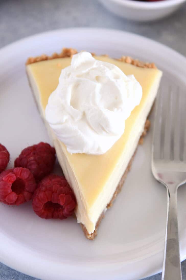Top-down view of a slice of lemon pie with a dollop of whipped cream on a white plate, with fresh raspberries on the side.