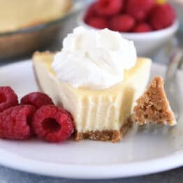 A slice of lemon pie with a graham cracker crust, topped with whipped cream and a bite taken out. Fresh raspberries sit beside the slice.