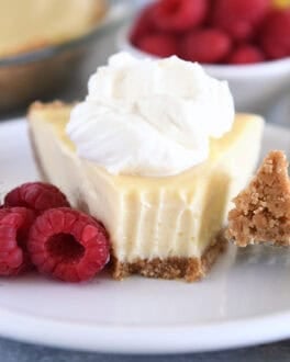 A slice of lemon pie with a graham cracker crust, topped with whipped cream and a bite taken out. Fresh raspberries sit beside the slice.