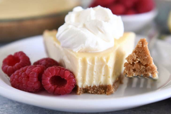 A slice of lemon pie with a graham cracker crust, topped with whipped cream and a bite taken out. Fresh raspberries sit beside the slice.