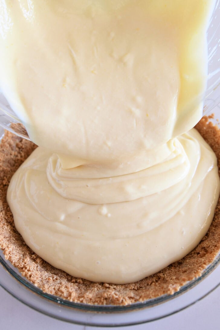 Lemon pie filling getting poured into a graham cracker crust in a glass pie pan.