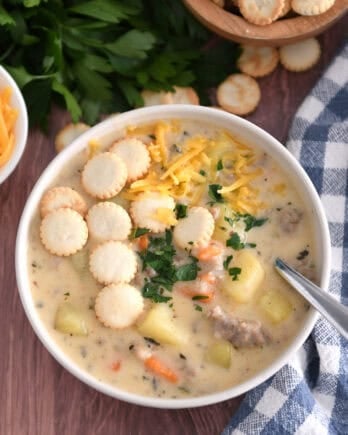 Soup bowl with sausage potato soup topped with oyster saltine crackers, fresh parsley and grated cheese and spoon on side.