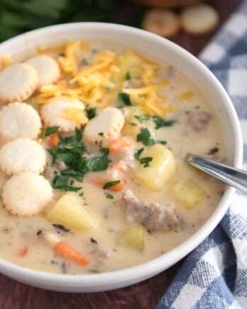 Soup bowl with sausage potato soup topped with oyster saltine crackers, fresh parsley and grated cheese and spoon on side.