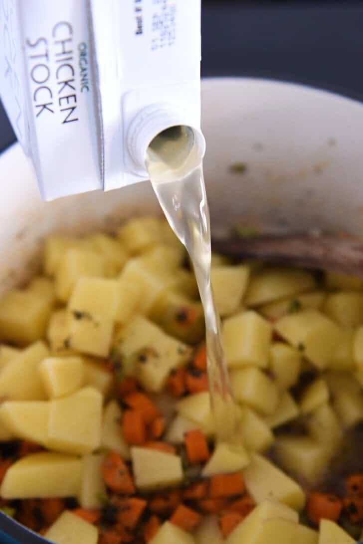 Pouring chicken stock into soup pot with potatoes and carrots.