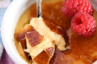 Spoon cracking through top shell of creme brûlée in white ramekin with raspberries on the side.