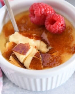Spoon cracking through top shell of creme brûlée in white ramekin with raspberries on the side.