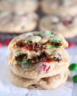 Two baked cookies with chocolate chips and red and green M&Ms with a cookie on top broken in half with melted chocolate chips.