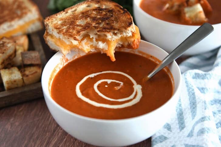 White bowl with homemade tomato soup, swirls of heavy cream, and half a grilled cheese sandwich balanced on side of bowl.