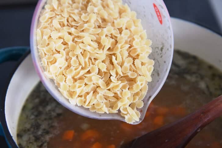 Small pasta in measuring cup over soup pot with broth and soup.