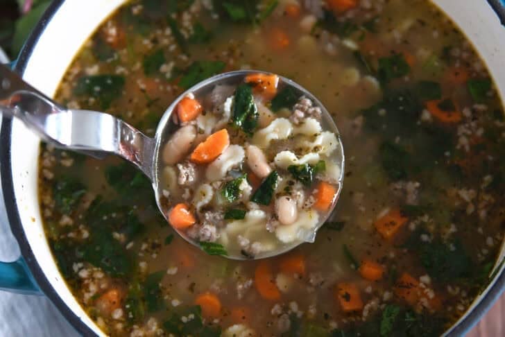 Ladle lifting serving of soup in pot with small pasta, spinach, carrots, white beans, and sausage.
