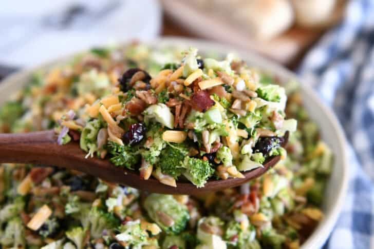 A wooden spoon taking a scoop of chopped broccoli salad with shredded cheese, diced onion, bacon bits, sunflower seeds, and dried cranberries coated in a light sauce.