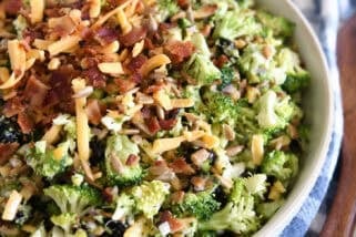 Top down view of a white bowl of chopped broccoli salad with shredded cheese, diced onion, bacon bits, sunflower seeds, and dried cranberries coated in a light sauce.