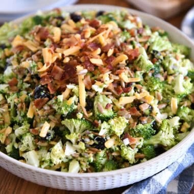 A white bowl of chopped broccoli salad with shredded cheese, diced onion, bacon bits, sunflower seeds, and dried cranberries coated in a light sauce.