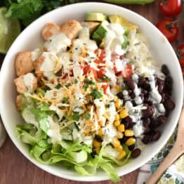 White bowl with cooked chicken, chopped tomatoes, cilantro lime rice, chopped avocados, black beans, roasted corn, and lettuce.