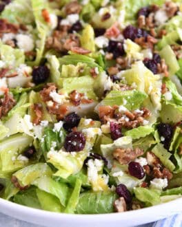 Top down view of a large white bowl full of chopped romaine salad with candied nuts, bacon bits, crumbled cheese, and dried cranberries.