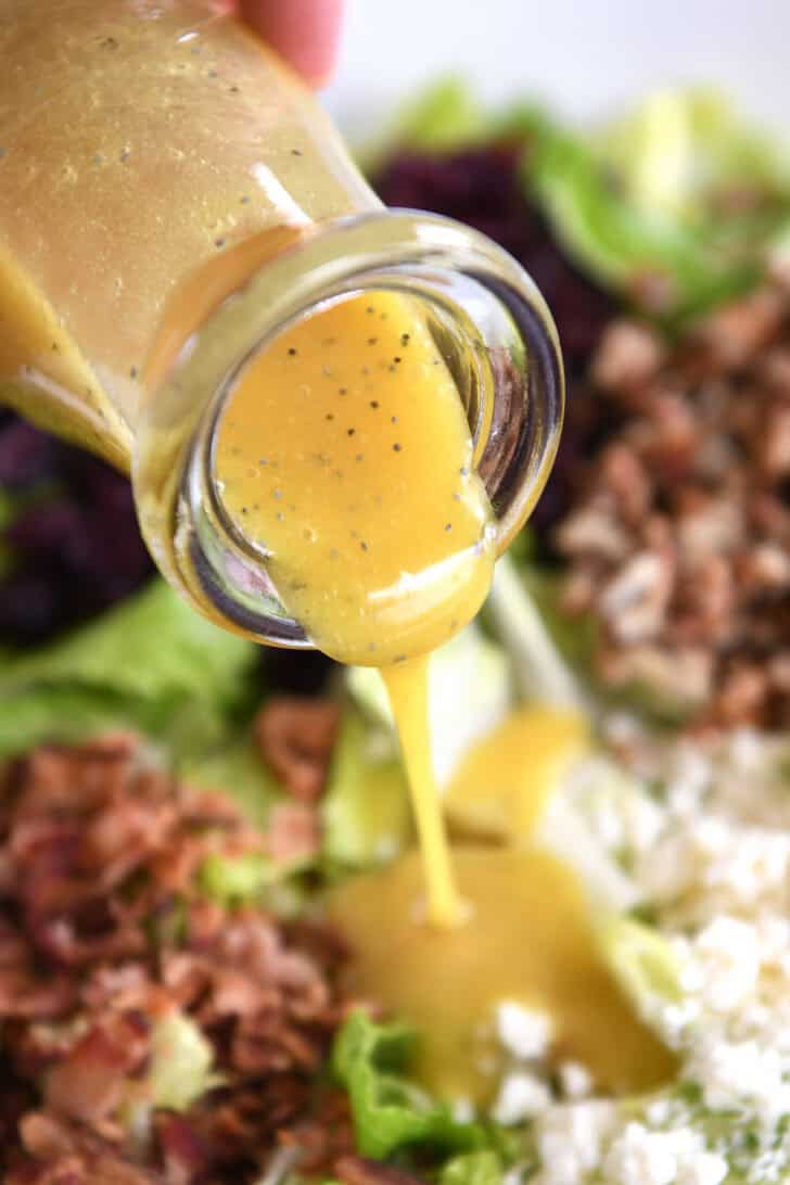 A glass jar of poppy seed dressing being poured over a chopped romaine salad.