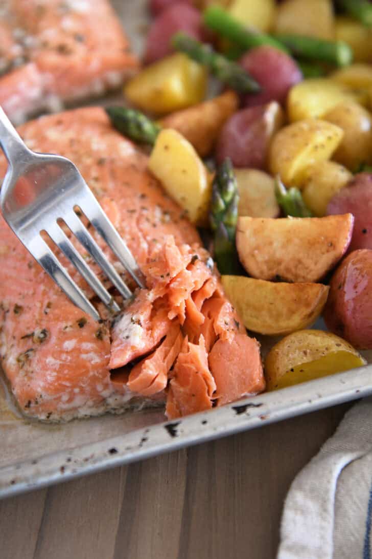 Fork flaking filet of baked salmon with roasted potatoes and asparagus on sheet pan.