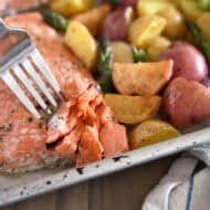 Fork flaking filet of baked salmon with roasted potatoes and asparagus on sheet pan.