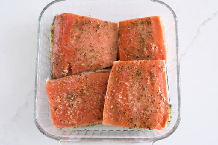 Top down view of glass square dish of four salmon filets marinaded in lemon juice, olive oil, and garlic.