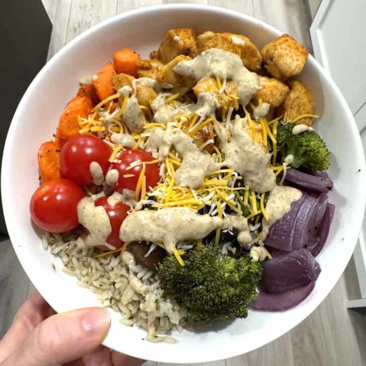 White bowl with cilantro lime rice, broccoli, onions, cherry tomatoes, carrots, chicken, cheese and Bitchin' sauce.