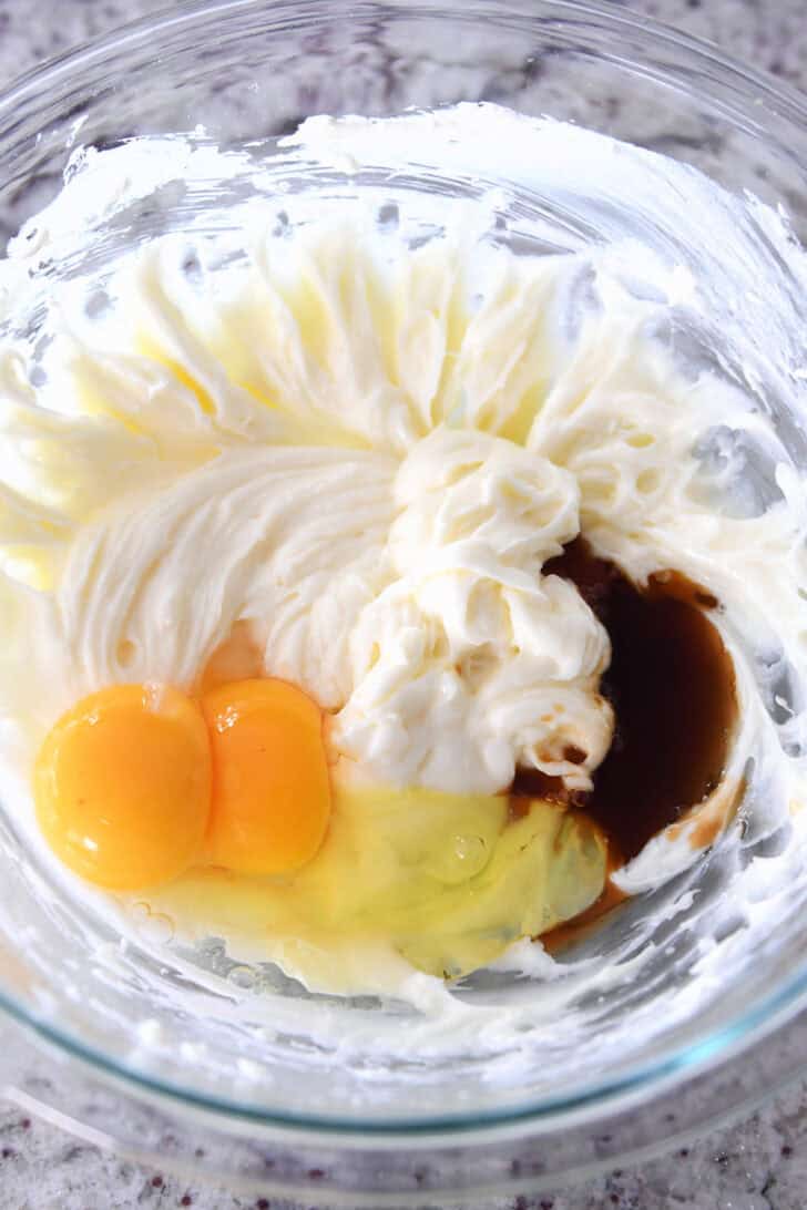 Top down view of a glass bowl with raw eggs, a cream cheese mixture, and vanilla.
