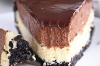 A white plate holding a slice of Oreo crust cheesecake, layered with creamy cheesecake, rich chocolate mousse, and topped with smooth chocolate ganache. A bite is taken out of the slice.