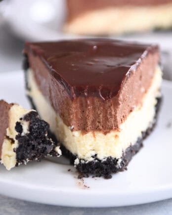 A white plate holding a slice of Oreo crust cheesecake, layered with creamy cheesecake, rich chocolate mousse, and topped with smooth chocolate ganache. A bite is taken out of the slice.