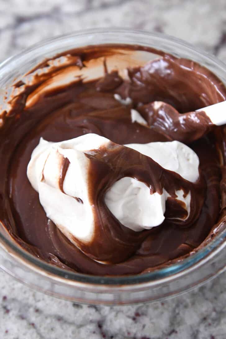 Top down view of whipped cream mixture getting stirred into a chocolate sauce with a rubber spatula.