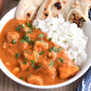 Top-down view of a white bowl with flatbread, white rice, and diced chicken in an orange Indian-style sauce, topped with fresh cilantro.