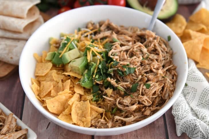 White bowl with salsa verde chicken, diced avocado, chopped cilantro, rice and tortilla chips.