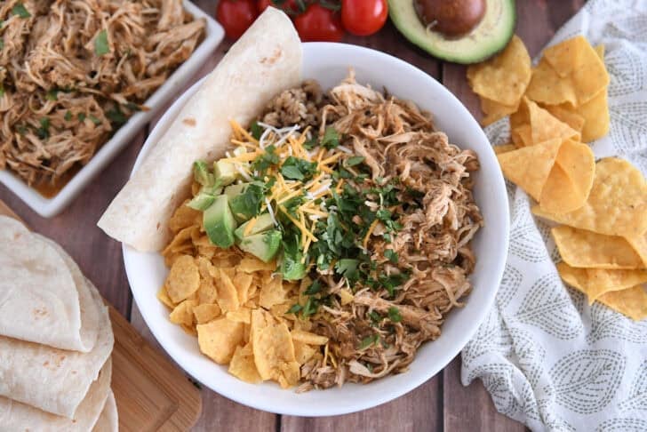 White bowl with salsa verde chicken, diced avocado, chopped cilantro, rice and tortilla chips.