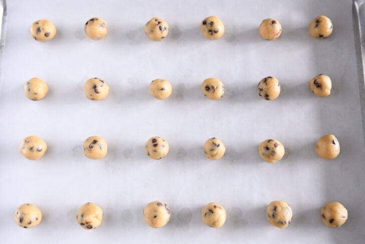 A baking tray containing 24 unbaked mini cookie dough balls is lined with parchment paper.