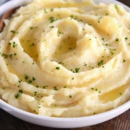 A white bowl with buttery mashed potatoes topped with parsley flakes. A wooden spoon is in the bowl.