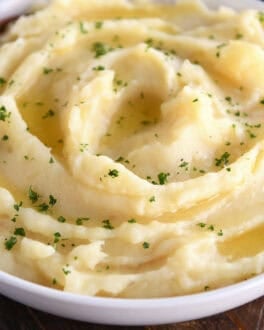 A white bowl with buttery mashed potatoes topped with parsley flakes. A wooden spoon is in the bowl.
