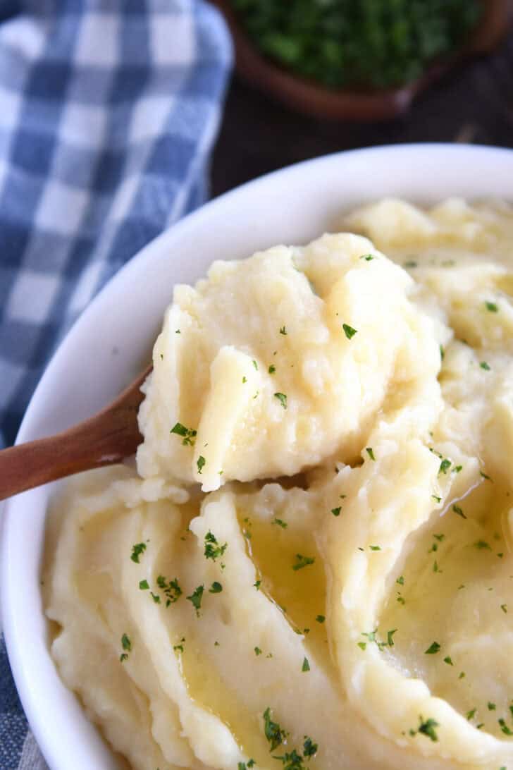 A wooden spoon taking a large scoop of buttery mashed potatoes out of a white bowl.