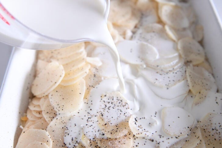 Pouring cream over thin slices of potatoes in white 9X13-inch dish.