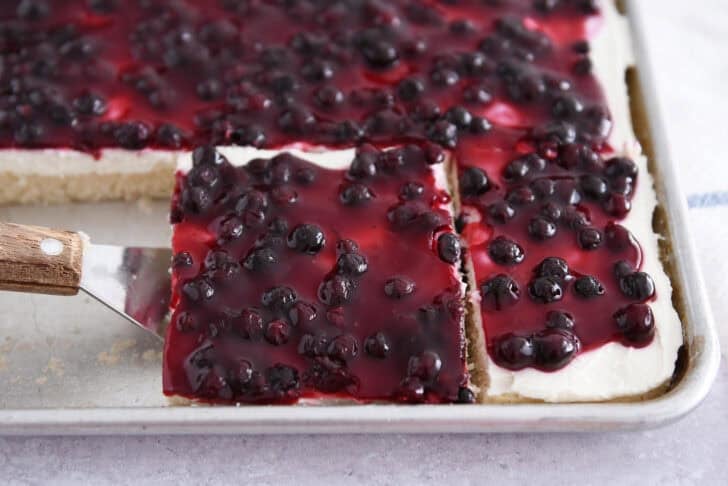 Square of white sheet cake topped with cream cheese layer and fruit topping on spatula.