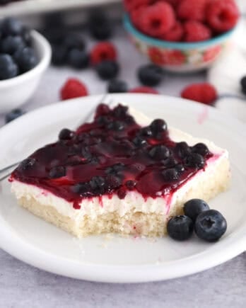 Square of white sheet cake topped with cream cheese layer and fruit topping on white plate with fresh blueberries.