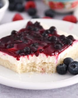 Square cake with white sheet cake topped with a layer of cream cheese and fruit topping on a white plate with fresh blueberries.
