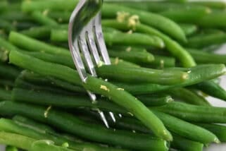 Fork piercing several fresh, cooked green beans on white tray.