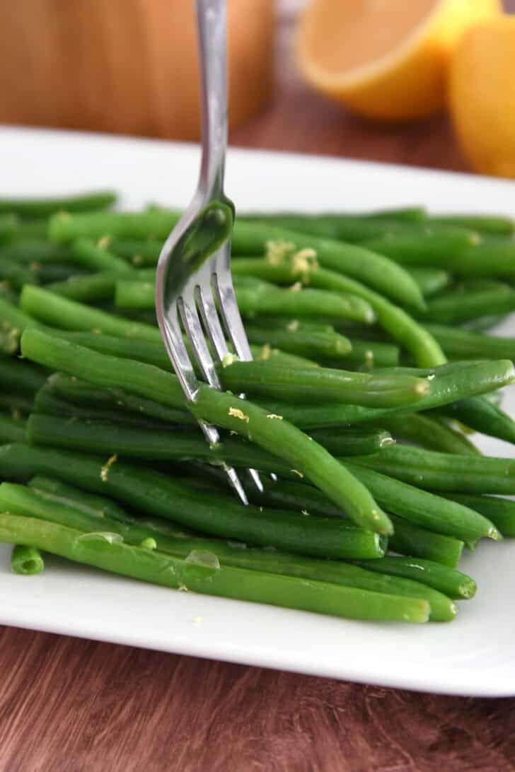 Fork piercing several fresh, cooked green beans on white tray.
