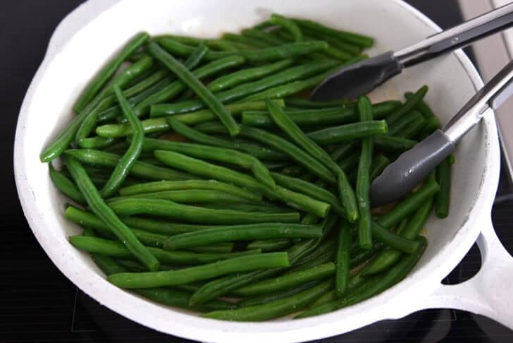 White skillet with cooked green beans, broth and tongs.