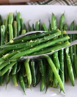 A white tray with cooked green beans, lemon zest and some scooped out green beans.