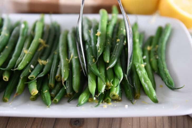 A white tray with cooked green beans, lemon zest and some scooped out green beans.