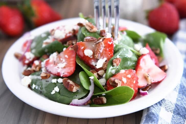 White plate with fresh spinach, sliced strawberries, red onions, pecans and poppy seed dressing.