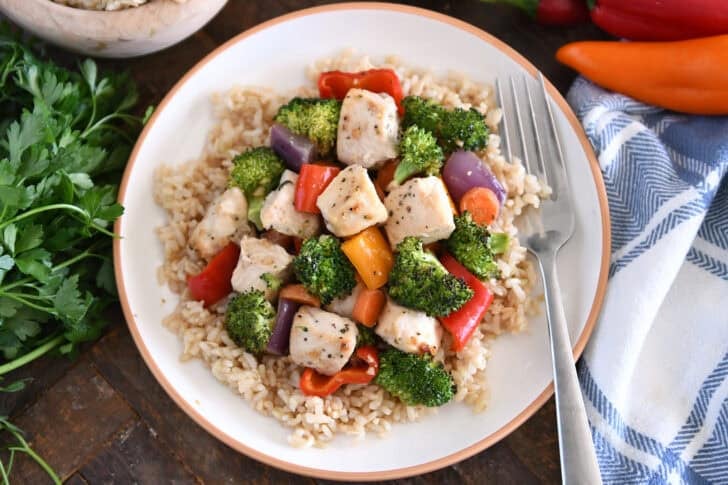 Serve brown rice, cooked chicken, broccoli, red onion, bell peppers, carrots and red onion on a white plate with tan rim.