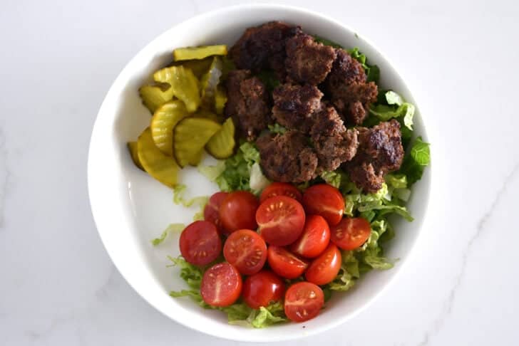 Top down view of white bowl with lettuce, ground beef, tomatoes and pickles.