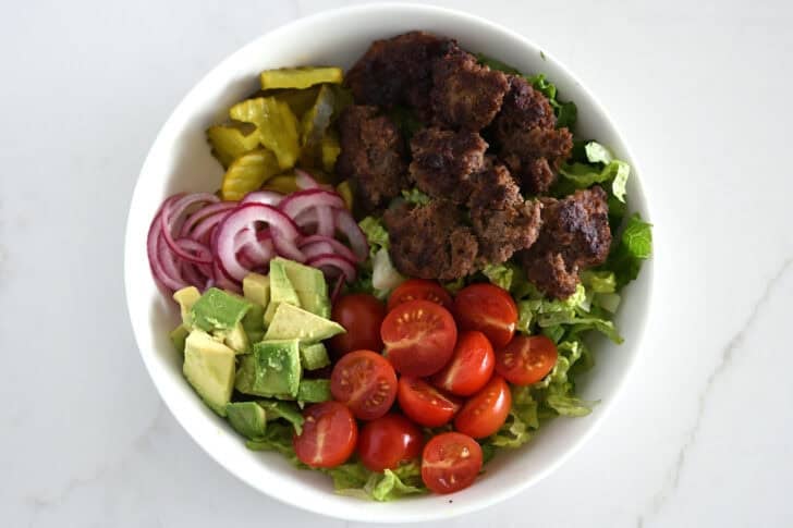 Top down view of lettuce, beef, tomatoes, avocado, pickled onions and pickles in white bowls.