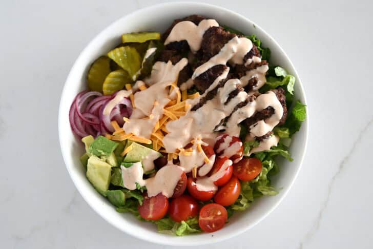 Top down view of lettuce, ground beef, tomatoes, avocado, pickled onions, pickles, shredded cheese and cream sauce in white bowl.